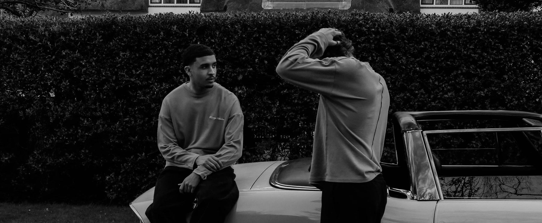 Two men sitting on a car in a driveway with a hedge in the background